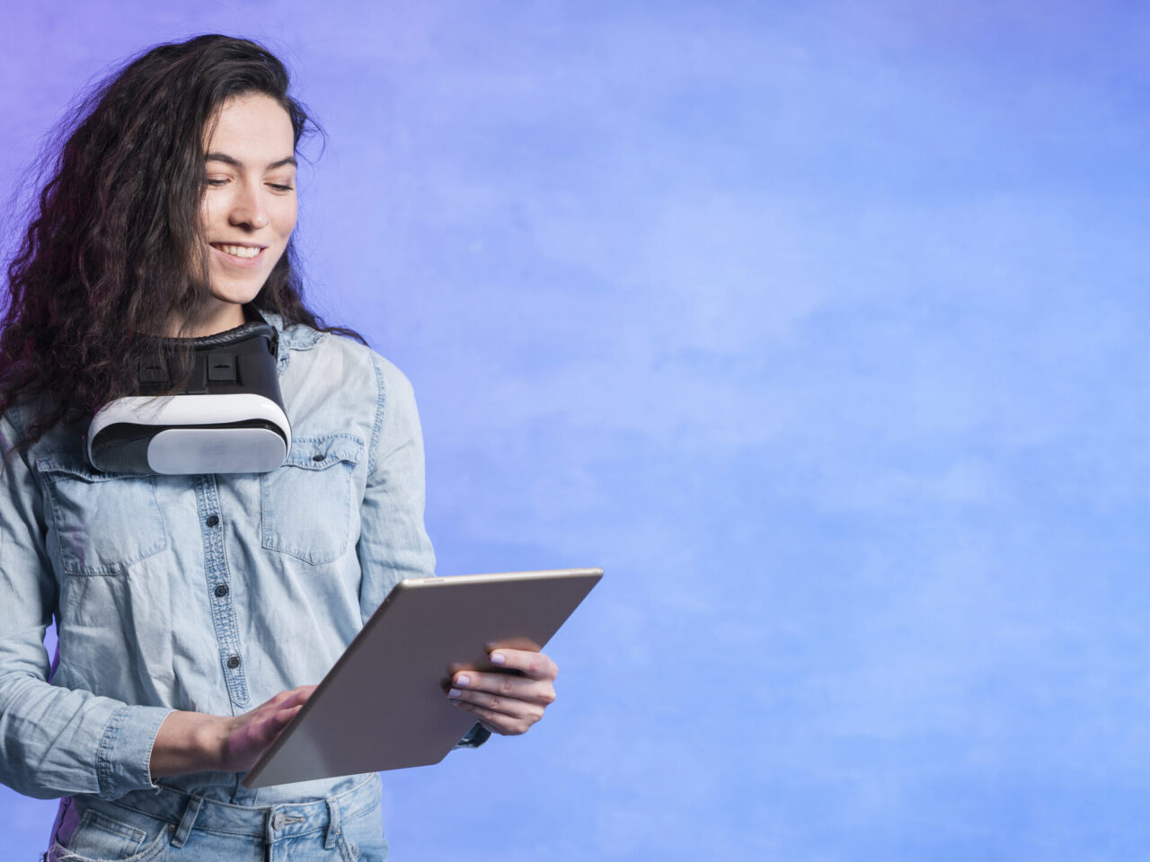 A young adult with curly hair wearing a denim shirt interacts with a tablet, a VR headset resting around their neck. The purple-blue gradient background evokes a sense of digital transformation and future readiness.