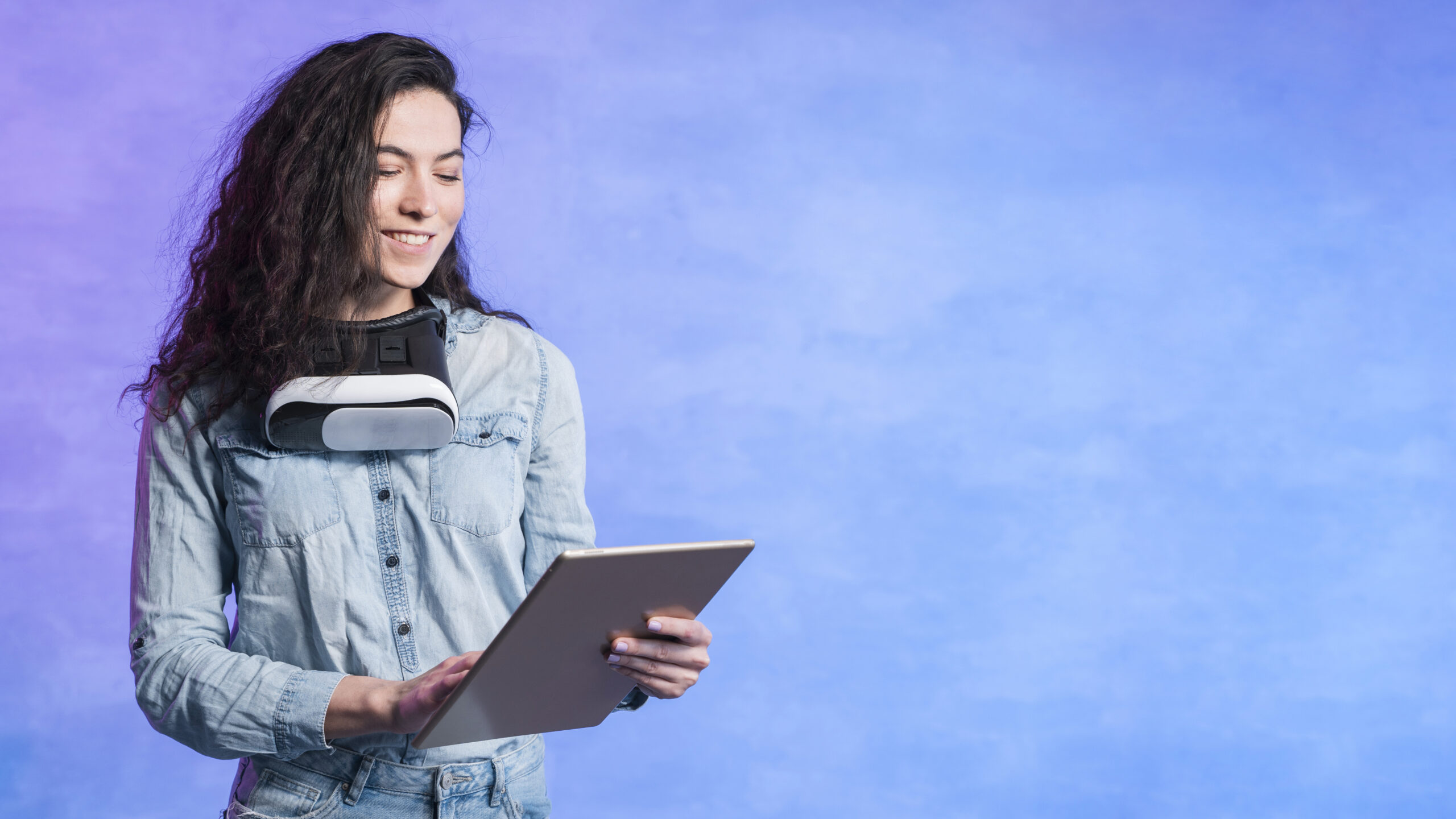 A young adult with curly hair wearing a denim shirt interacts with a tablet, a VR headset resting around their neck. The purple-blue gradient background evokes a sense of digital transformation and future readiness.