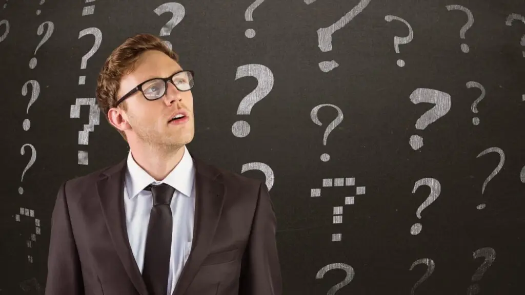 A man in a suit stands before a blackboard filled with scattered white question marks. His upward gaze and contemplative expression capture the uncertainty students often feel when choosing a university program.