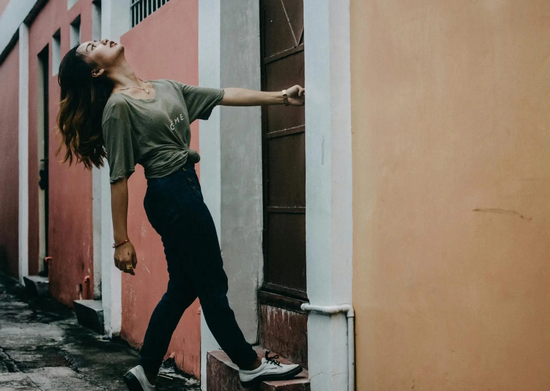 A young person in a green t-shirt leans dramatically on a narrow urban sidewalk, framed by pastel-colored walls. Their pose and motion suggest boldness, self-expression, and the journey of discovering one’s true identity.