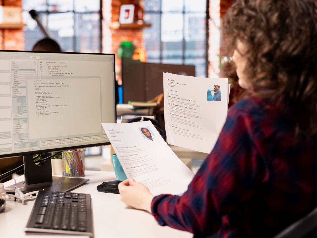 Recruiter reviewing printed resumes at a desk while comparing candidate profiles on a computer screen.